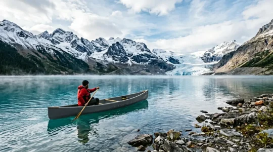 A solo canoeist navigating turquoise glacial lake waters surrounded by snow-capped peaks under dramatic lighting