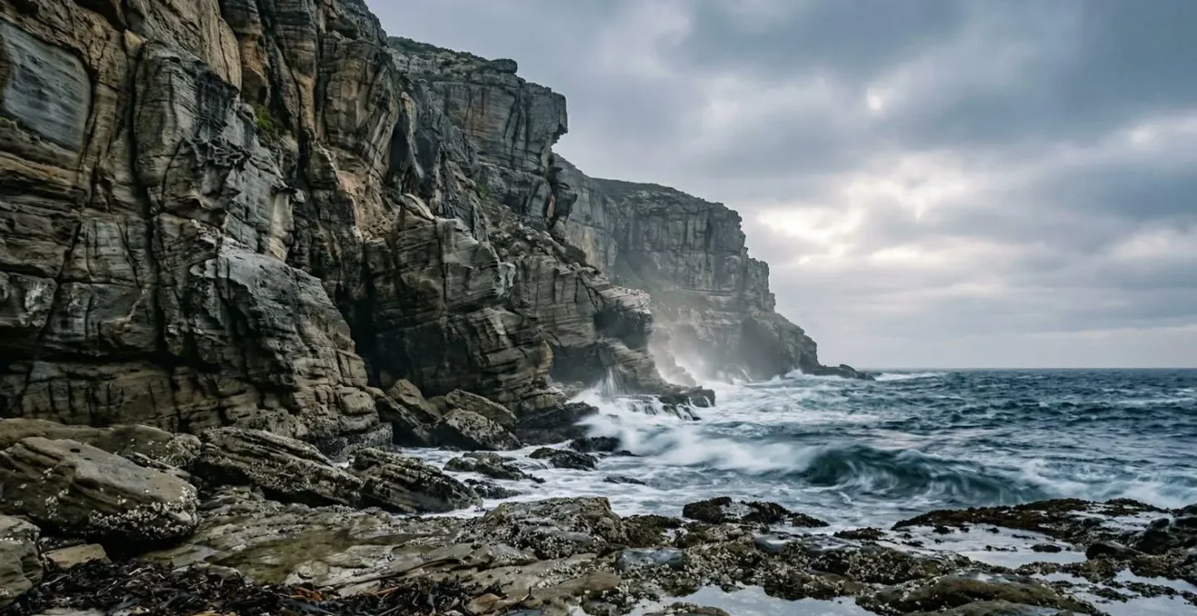 Dramatic coastal cliff with jagged rocks and incoming tide creating spray against weathered sedimentary walls
