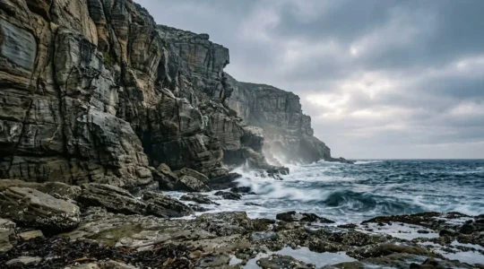 Dramatic coastal cliff with jagged rocks and incoming tide creating spray against weathered sedimentary walls