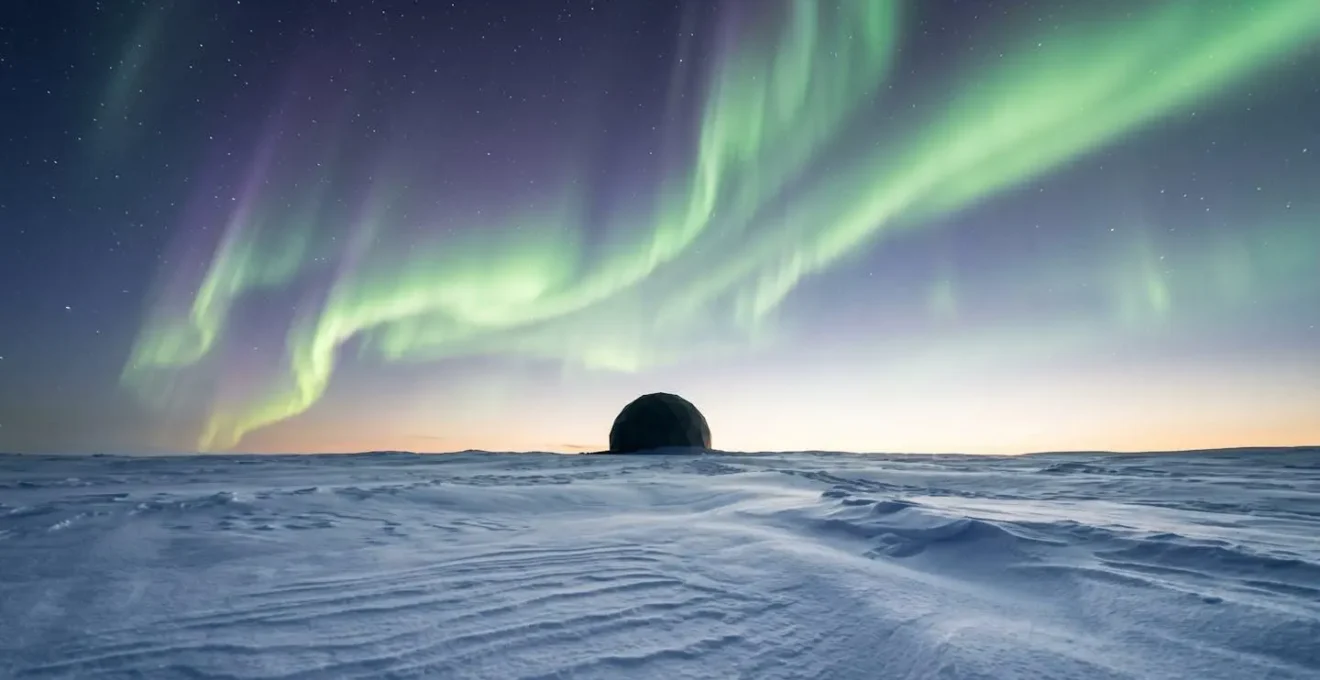 Wide-angle environmental photograph of Arctic landscape under aurora borealis with minimalist glass dome structure in distance
